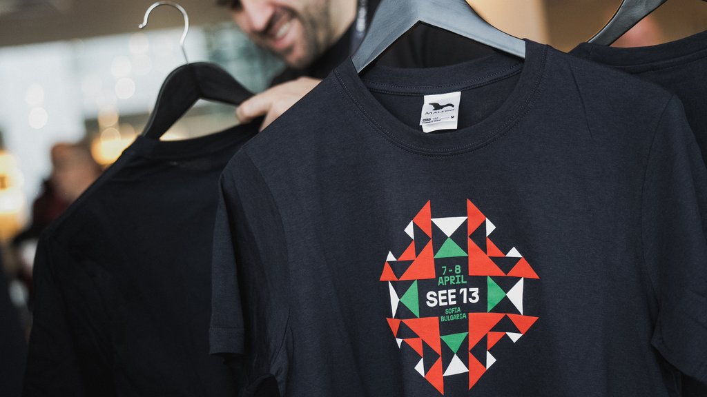 SEeE 13 t shirts hang on black hangers and a man in the background has picked one up and smiled.