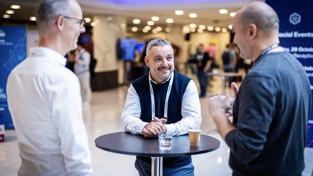 Three men stand around a tall table talking.