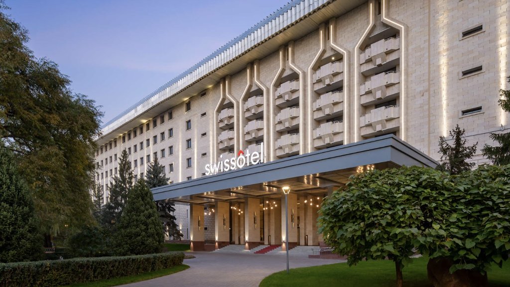 A photo of a Swissôtel property, showing a grand exterior with geometric architecture and warm lighting. The building has a modernist vibe with a clean facade, rows of balconies, and nicely manicured greenery leading up to the main entrance.